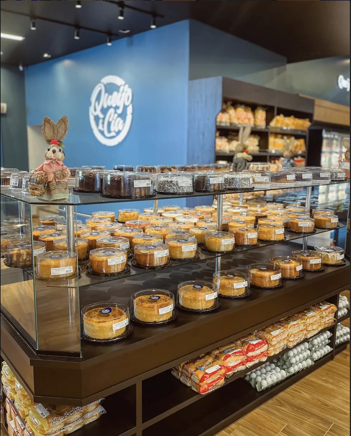 Interior da loja Queijo & Cia: balcão refrigerado com bolos e tortas, e a parede azul com o logo da marca ao fundo