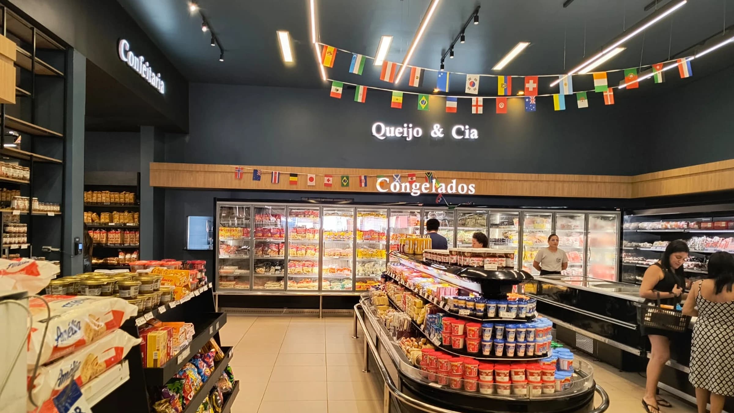 Corredor da loja Queijo & Cia, com o balcão de Congelados e o letreiro iluminado Queijo & Cia na parede